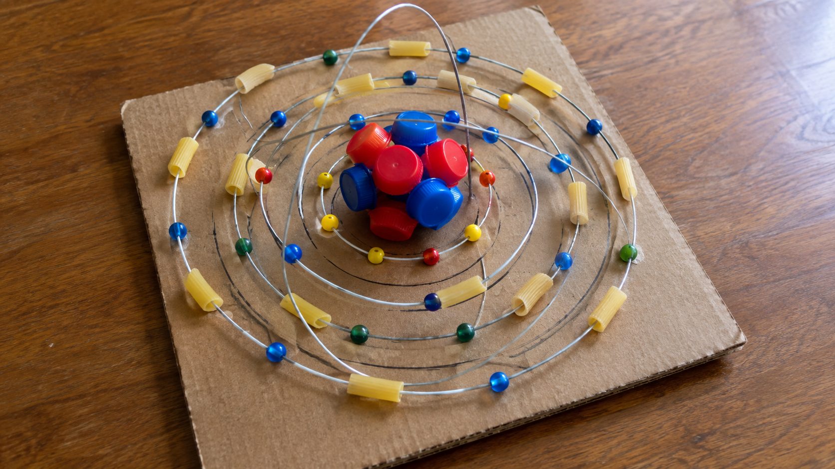 Recycled material 3D atom model built from bottle caps, beads, and aluminum wire with shells drawn on a cardboard base